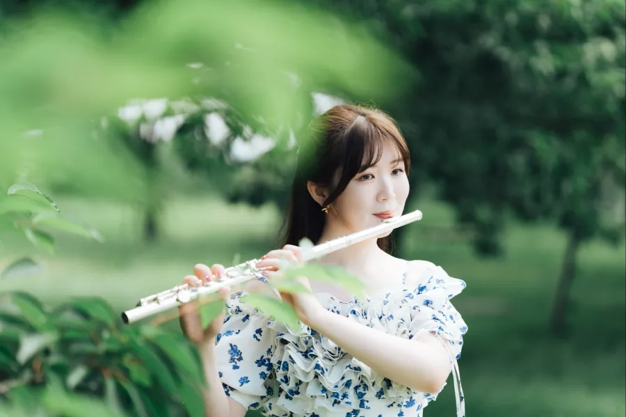 森の中で、白地に青い花柄のドレスを着た笑顔の永田梨紗がフルートを吹いている写真